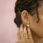 A high-resolution studio shot of the Mariposa 14K Yellow Gold Heart Lab Grown Diamond Earring Jackets, styled on a female model with a soft, pastel pink background that evokes Valentine's Day romance. The earrings are subtly displayed on the model’s ears, 