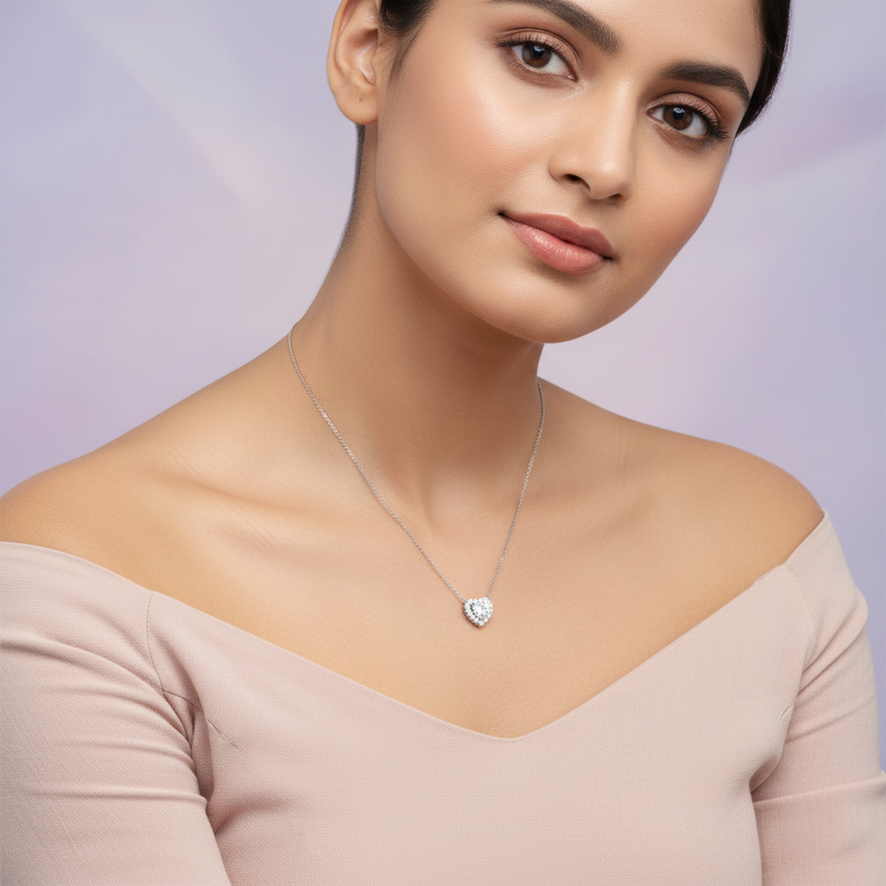 A female model in a gentle, natural pose, seated against a dreamy pastel purple background that evokes a romantic, feminine atmosphere. She wears a light, off-shoulder blouse that subtly complements the necklace. The Valentina heart pendant is clearly visi