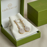 Close-up of Ambery 14K yellow gold diamond drop earrings in packaging, highlighting gold setting and sparkling diamonds with soft natural light.