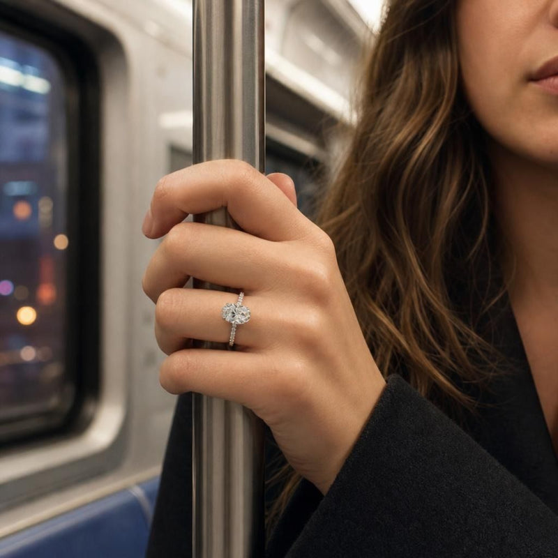 Close-up of Adriana engagement ring on hand with city lights in background, showcasing the oval lab-grown diamond and hidden halo.