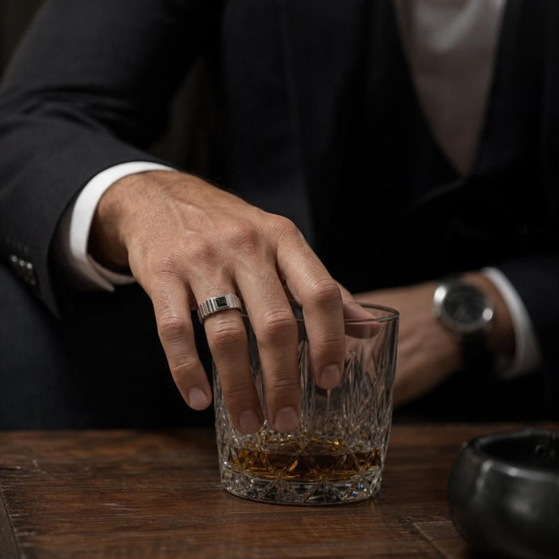 Princess-cut black spinel solitaire ring in 925 sterling silver worn on a man's hand resting over a whiskey glass.
