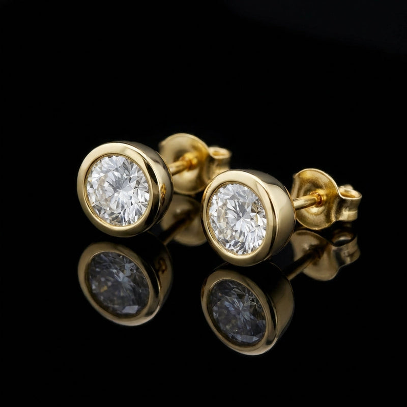 A close-up studio shot of the Alisha 1.5 Ct TW Non Certified Lab Grown Round Diamond Studs Bezel 14K Yellow Gold Earrings, photographed with a softbox lighting setup that highlights the brilliance of the diamonds and the warm tone of the gold. The camera i