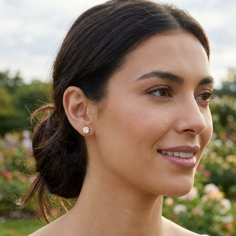 A lifestyle scene featuring a human model wearing the Alisha diamond earrings, set against a softly blurred outdoor garden backdrop bathed in natural diffused daylight. The model’s head is turned slightly to showcase the earrings on the earlobe, with gentl