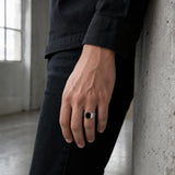 Compact black spinel solitaire ring on a man's finger against an industrial concrete pillar.